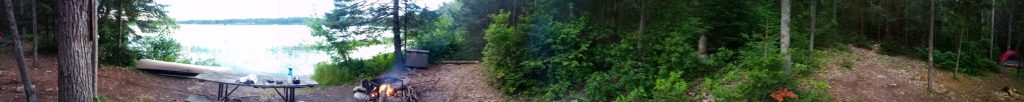 First panoramic photo of campsite B4 on Lake Pakeshkag at Grundy Lake Provincial Park. Lake, canoe fire pit, food locker, and picnic table on the left. Woods in the center and tent is far off on the right.