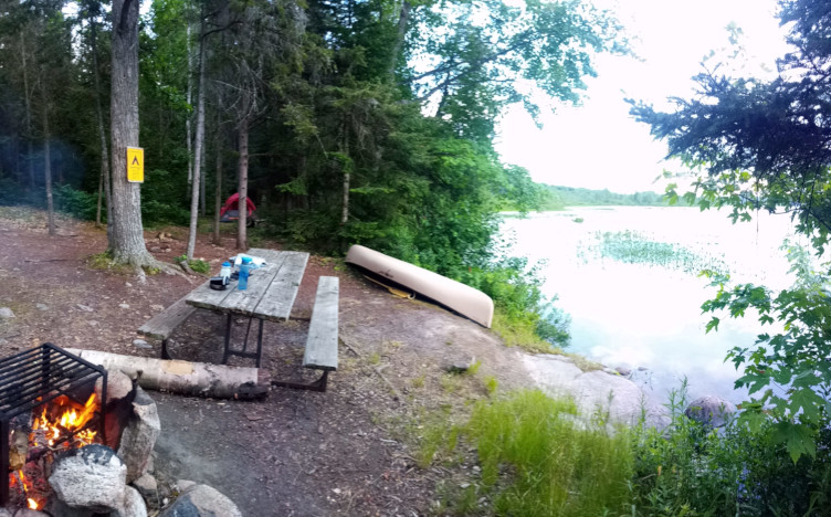 View of campsite with the lake on the right, the picnic table and canoe in the center with the tent far in the background. On the left foreground is the fire pit with fire.