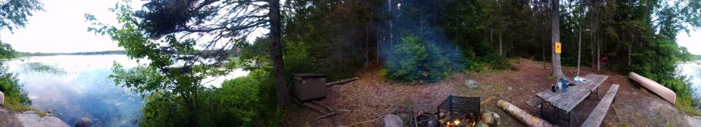Second panoramic photo of campsite B4 on Lake Pakeshkag at Grundy Lake Provincial Park. Lake on the left. Food locker and fire pit in the center. On the right is the picnic table, canoe and far in the distance is the tent.