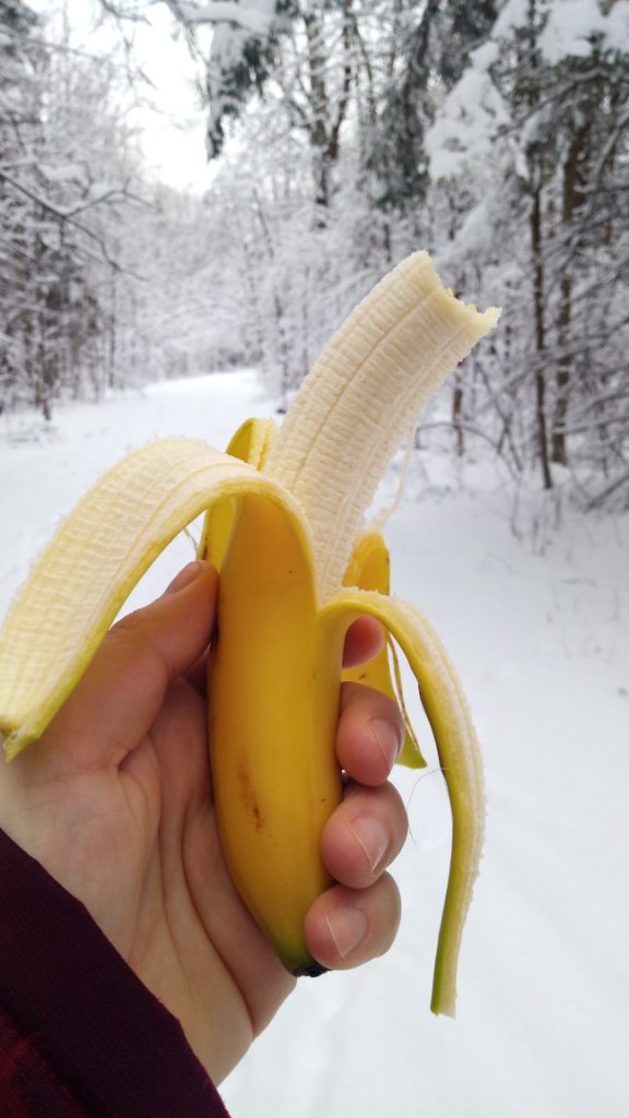 Holding a half peeled banana with a bite taken out and a snowy trail in the background